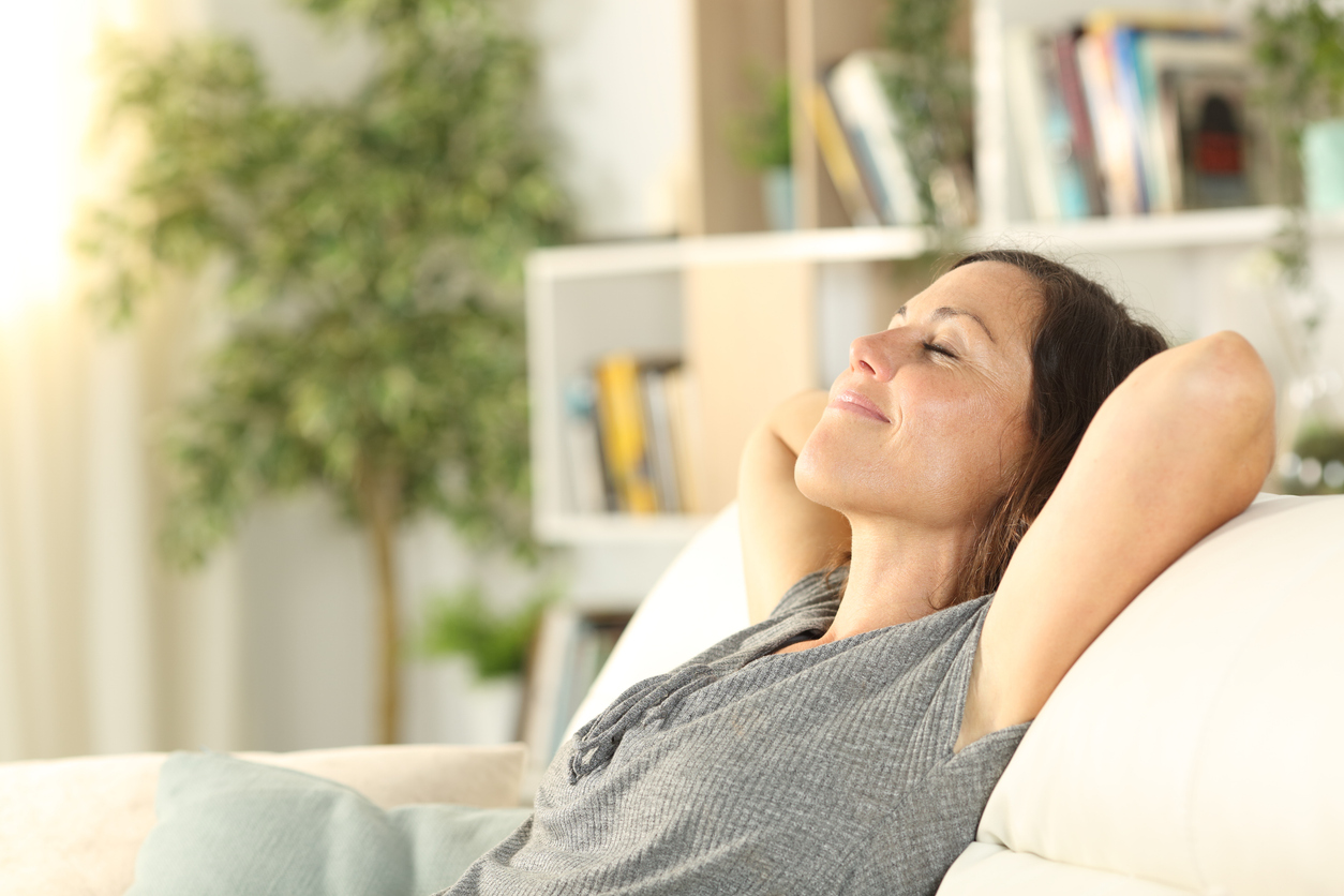 Woman breathes easy and relaxes at home, sitting on the couch with her arms behind her head.